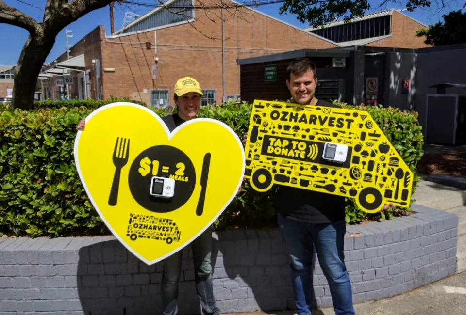 Two OzHarvest volunteers holding cardboard cut outs of a love heart and a ozharvest van $1 two meals